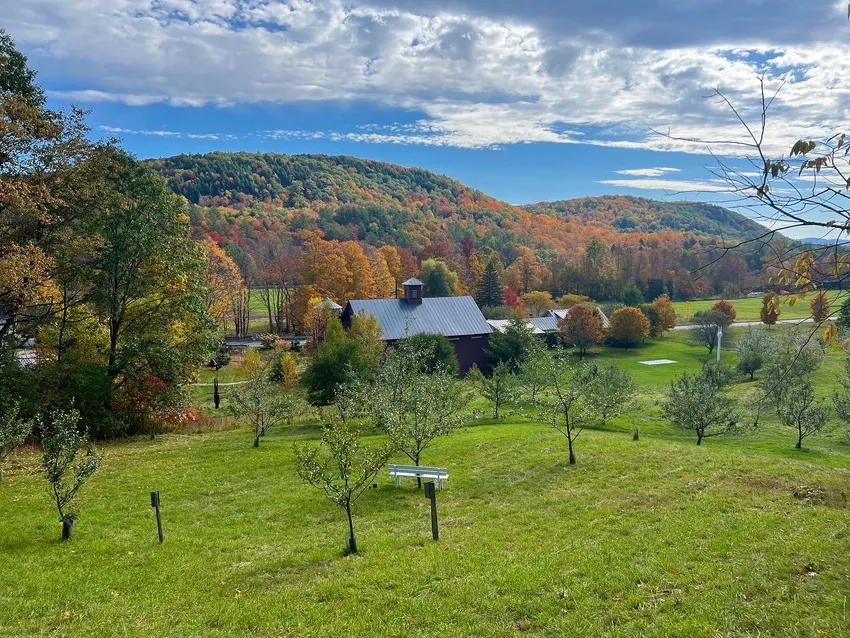 “Autumn view across the Hall Art Foundation campus in Reading, Vermont, with a dark barn surrounded by green fields, orchard trees, and colorful wooded hills beneath a blue and cloudy sky.