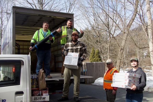 Five Home Depot volunteers unload flooring and painting supplies from a truck outdoors.